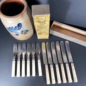 May include: An antique stoneware crock with blue floral designs, a box of "AMERICAN TABLE CUTLERY CO." knives and forks, and a set of vintage silverware. The silverware includes forks and knives with white handles.
