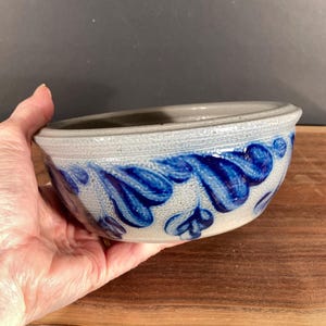 May include: A stoneware bowl with a light gray base and a blue floral design. The bowl has a textured rim and is held in a hand. The blue design is painted in a stylized leaf pattern.