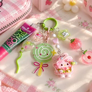 Puede incluir: Un tubo de brillo labial Essence Juicy Bomb junto a un llavero verde con un dije de piruleta verde, un dije de Hello Kitty rosa, una estrella verde, una cuenta de fresa roja y una cuenta con forma de melocotón rosa en un anillo verde.