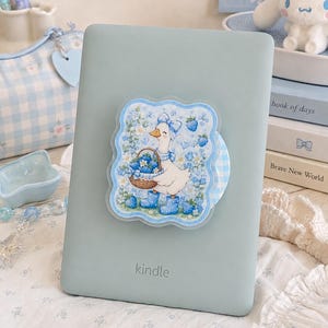 May include: A light sage green Kindle e-reader featuring a decorative sticker on the back. The sticker displays a white goose wearing a blue bow and blue rain boots, holding a basket of blue strawberries amidst a background of blue flowers.