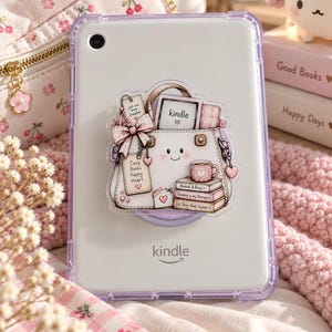 Puede incluir: Una funda protectora transparente para un lector electrónico Kindle con una pegatina de un bolso blanco con cara sonriente. El bolso contiene un Kindle, una taza de café y una pila de libros con texto como "Cozy Books Happy Heart" y "Booked & Busy".