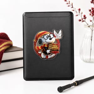 May include: A black rectangular e-reader featuring a circular sticker of a white cartoon dog wearing round glasses, a red and yellow striped hat, and a matching scarf. The dog holds a stack of books, one labeled MAGIC, with a small white owl nearby.
