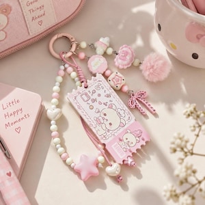 Op de afbeelding: Een roze sleutelhanger met een rechthoekige ticketvormige bedel met een berenillustratie, omringd door een kralenlus met hart- en stervormige bedels, een pluizige pompon en een kleine strik bevestigd aan een ronde metalen ring.