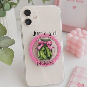 Könnte beinhalten: Eine transparente Smartphone-Hülle mit einem runden, pinken Handyhalter. Das Design zeigt eine Grafik eines grünen Gurkenglases mit einer pinken Schleife und dem schwarzen Text: just a girl who loves pickles. Die Hülle hat einen Ausschnitt für die Doppelkamera.