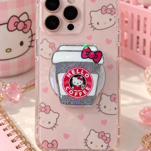 Può includere: Una custodia trasparente per smartphone con un motivo ripetuto di volti di Hello Kitty e piccoli cuori rosa. Al centro è fissato un supporto per telefono glitterato a forma di tazza di caffè con un logo circolare che recita HELLO COFFEE.