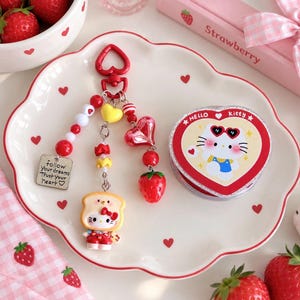 Può includere: Un portachiavi rosso a forma di cuore con una figura di Hello Kitty vestita da toast, un ciondolo a forma di fragola e un ciondolo quadrato argentato con il testo follow your dreams Trust your heart, accanto a un contenitore Hello Kitty a cuore.
