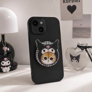 Op de afbeelding: Een zwarte smartphonehoes met een ronde telefoonhouder. De houder toont een afbeelding van een rode kat met een zwarte capuchon met een roze schedel-embleem, tegen een achtergrond met een zwart-wit geblokt patroon.