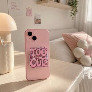 Puede incluir: Una funda de smartphone de color rosa claro con un soporte circular en la parte posterior. El accesorio muestra el texto "TOO CUTE" en letras grandes de color rosa estilo burbuja, decorado con corazones, un lazo pequeño y destellos.