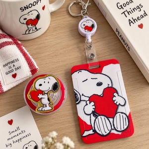 May include: A white ceramic mug, a retractable badge reel with a Snoopy and heart graphic, and a vertical ID card holder featuring a large Snoopy illustration holding a red heart. The items include text such as SNOOPY, Good Things Ahead, and Happiness is a Good Day.