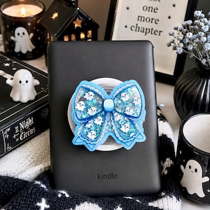 Puede incluir: Un lector electrónico Kindle negro con un soporte para teléfono en forma de lazo azul adherido a la parte posterior. El lazo está relleno de líquido azul, purpurina y pequeñas figuras de fantasmas blancos. Tiene un borde festoneado y un centro azul.