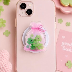 Può includere: Uno smartphone rosa chiaro con un supporto trasparente applicato sul retro. Il supporto presenta un design a sacchetto trasparente contenente diversi piccoli quadrifogli verdi sorridenti, legati in cima con un cordino rosa acceso.