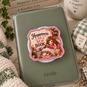Op de afbeelding: Een saliegroene e-reader met een vierkante, glitterende zelfklevende telefoonhouder. De houder toont een illustratie van een persoon die een boek leest met een kat en een teddybeer, met de tekst "Happiness is a NEW BOOK" in een decoratief kader.