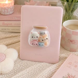 Puede incluir: Un lector electrónico Kindle de color rosa claro con un agarre adhesivo transparente en forma de frasco en la parte posterior. El frasco contiene cinco pequeñas figuras de gatos estilo caricatura en blanco, naranja, rosa, azul y gris.