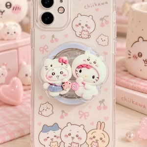 May include: A clear smartphone case featuring a pattern of small pink hearts, cherries, and cartoon characters with the text chiikawa. A circular phone grip is attached to the centre, decorated with two 3D figures of Hello Kitty wearing character hoods.