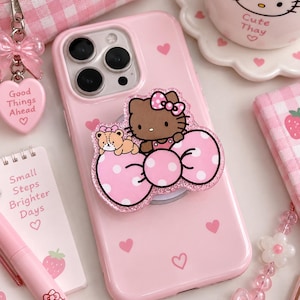 Puede incluir: Una funda de smartphone rosa claro con un accesorio brillante de Hello Kitty que incluye un gran lazo rosa de lunares y un pequeño oso. La funda está decorada con pequeños corazones rosas y diseñada para un dispositivo con triple cámara.