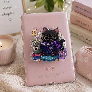 Può includere: Un lettore e-book Kindle rosa chiaro con una custodia protettiva trasparente e un adesivo di un gatto nero con occhiali e sciarpa viola, che legge un libro con la scritta GOOD VIBES, seduto su un cuscino blu a pois.