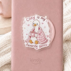 May include: A dusty rose Kindle e-reader featuring a clear sticker of a white duck wrapped in a pink plaid blanket, holding a pink mug. The sticker text reads, "Cozy is my favourite genre," surrounded by small pink hearts and gold star accents.