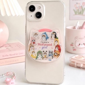 May include: A clear smartphone case featuring a cat-shaped pop-out grip. The grip displays multiple illustrations of small cats dressed in various costumes, including a shark, a burger, and fruit, with the text I'm just a Mofusand girl.