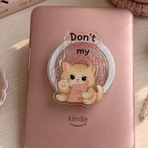 Op de afbeelding: Een roségouden Kindle e-reader met een ronde telefoonhouder aan de achterkant. De houder toont een cartoonillustratie van een pluizig oranje kitten dat een kleine tablet vasthoudt met de tekst Don't touch my Kindle erboven gedrukt.