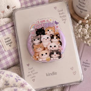 May include: A clear Kindle case featuring a circular phone grip with a graphic of nine cartoon cats and the text 'i am a Cat girl'. The case includes printed text: 'Good Books Happy Days', 'Just a girl who loves cats', and 'A Brighter Chapter' with hearts.