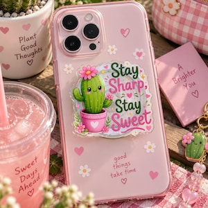 May include: A clear phone case featuring a pink tint and a circular grip attachment. The grip displays a green cactus in a pink pot with the text "Stay Sharp, Stay Sweet". The case is decorated with small white flowers, pink hearts, and the text "good things take time".