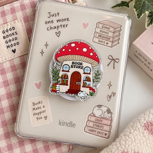 Op de afbeelding: Een doorzichtig Kindle-hoesje versierd met diverse stickers in boekenthema en een centrale glitterende telefoonhouder in de vorm van een paddenstoel. De stickers bevatten teksten zoals Just one more chapter, Good books brighter days en Books make a happier you.
