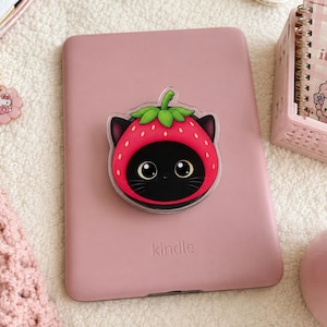 Puede incluir: Un soporte circular para teléfono con un gato negro que lleva una capucha roja en forma de fresa con hojas verdes. El gato tiene ojos grandes y está fijado en el centro de un lector electrónico rosa claro con la palabra kindle impresa.