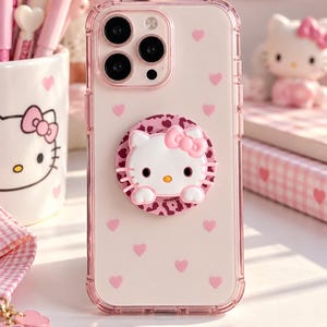 Puede incluir: Una funda transparente para smartphone con borde rosa y pequeños corazones rosas. La funda incluye un soporte circular decorado con estampado de leopardo en tonos rosa y un personaje de gato blanco en relieve con un lazo de color rosa.