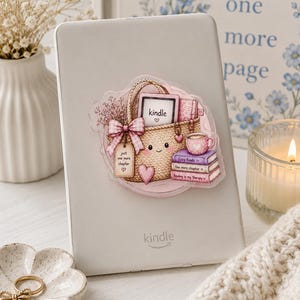 Può includere: Un lettore e-book bianco con un adesivo raffigurante un cesto intrecciato pieno di libri, un dispositivo Kindle, una tazza rosa e un fiocco rosa. L'adesivo include il testo "just one more chapter", "Cory Reads", "One more chapter" e "Reading is my therapy".