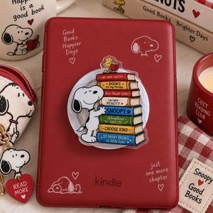 Puede incluir: Un lector electrónico Kindle rojo con una pegatina circular de Snoopy abrazando una pila de libros coloridos. El dispositivo tiene dibujos de líneas blancas de Snoopy y el texto Good Books Happier Days y just one more chapter con corazones.