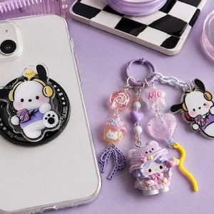 Puede incluir: Una funda transparente para smartphone con un soporte negro circular que muestra un perro de dibujos animados con auriculares amarillos. Al lado hay un llavero morado con cuentas, un dije de cupcake, un corazón y un personaje de gato.