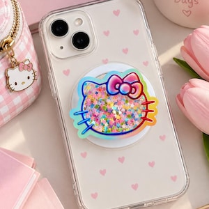 May include: A clear smartphone case featuring a repeating pattern of small pink hearts and a circular phone grip attachment. The grip displays a holographic outline of a cat character with a bow, filled with multicoloured star-shaped glitter.