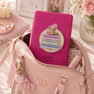 May include: A bright pink Kindle e-reader featuring a sticker of a worm wearing glasses and a pink hat reading a stack of books labelled with genres like fantasy and mystery, placed inside a light pink handbag with a heart-shaped metal emblem.