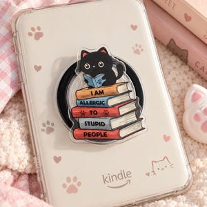 Op de afbeelding: Een doorzichtige Kindle-hoes met een zwarte kat die op een stapel boeken leest met de tekst I AM ALLERGIC TO STUPID PEOPLE. De hoes is versierd met kleine roze pootafdrukken en hartjes en bevat een zwarte ronde houder voor extra grip.