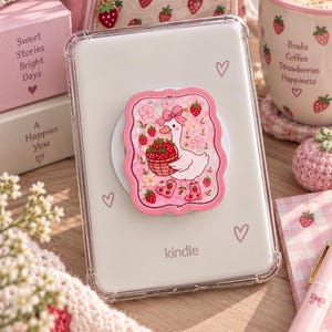 以下が含まれることがあります： 透明なKindleケースと、ピンク色の長方形のシェイカー型スマホグリップ。グリップには、ピンクのリボンとイチゴ柄のブーツを身につけ、イチゴの入ったバスケットを運ぶ白いアヒルのキャラクターが描かれており、周囲にはピンクの花とラメが散りばめられています。