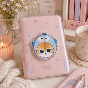 Op de afbeelding: Een lichtroze Kindle e-reader in een doorzichtige beschermhoes met iriserende glitteraccenten. Een ronde houder is in het midden bevestigd, met een afbeelding van een kat die een blauw kostuum met capuchon en grote ogen draagt.