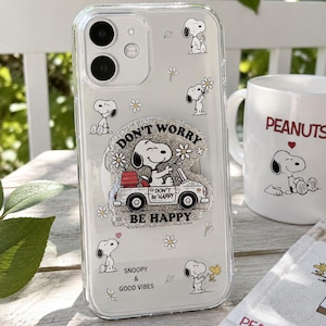 May include: A clear smartphone case featuring a Snoopy graphic pattern with small white daisies. A central glittery circular phone grip displays Snoopy driving a car with the text DON'T WORRY BE HAPPY. The case also includes the text SNOOPY & GOOD VIBES.
