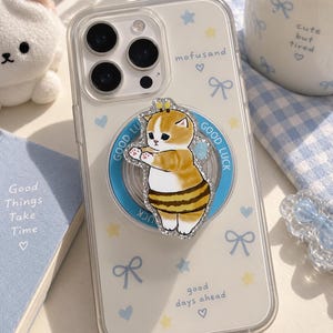 Puede incluir: Una funda transparente para smartphone con un patrón de lazos azul claro, estrellas amarillas y corazones pequeños. Incluye un soporte circular con borde azul y el texto GOOD LUCK, rematado con una ilustración brillante de un gato disfrazado de abeja.