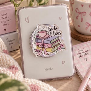 Può includere: Un lettore e-book Kindle bianco in una custodia protettiva trasparente con un supporto adesivo circolare. Il supporto mostra un'illustrazione di una pila di tre libri viola, rosa e gialli, sormontata da una tazza rosa e dal testo Books & Bliss.
