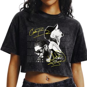 Könnte beinhalten: Ein schwarzes, säuregewaschenes bauchfreies T-Shirt mit einem schwarz-weißen Grafikdruck von zwei Personen. Das Design enthält den wiederholten Text "RAYMOND BROWN" in Weiß und zwei gelbe, handschriftliche Signaturen über dem Bild.