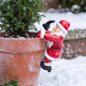 May include: A terracotta flower pot containing a small evergreen shrub, featuring a decorative Santa Claus figurine climbing the side. The Santa figure wears a red suit with white trim and black boots, and the pot is dusted with a light layer of snow.