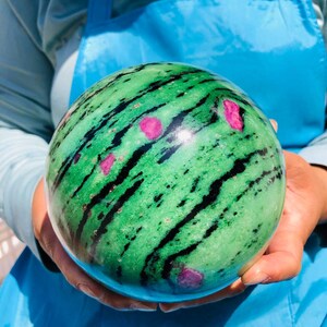 May include: A polished spherical stone featuring a vibrant green base with prominent black streaks running diagonally across the surface, accented by several small, distinct bright pink inclusions scattered throughout the round mineral specimen.