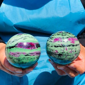 May include: Two polished spherical stones featuring a vibrant mix of light green zoisite, deep magenta ruby inclusions, and thin black hornblende streaks. Each smooth, round mineral specimen displays a unique natural pattern of contrasting colors.