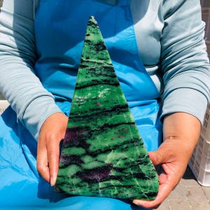 May include: A triangular-shaped polished stone slab featuring a vibrant green base with prominent black mineral veins and scattered deep magenta-colored inclusions throughout the surface, displaying a smooth texture and natural crystalline patterns.