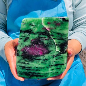 May include: A large, polished slab of zoisite mineral featuring a vibrant green base color, prominent black hornblende inclusions, and distinct patches of deep magenta ruby crystals throughout the stone's textured, irregular surface.