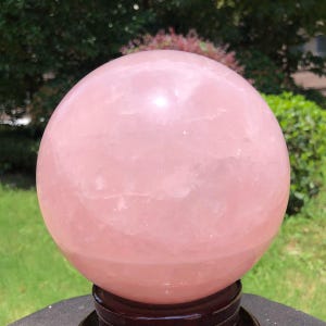 May include: A large, smooth, spherical rose quartz crystal with a translucent, pale pink hue and visible internal crystalline structures. The polished stone rests securely on a dark, circular wooden stand, showcasing its natural light reflection.