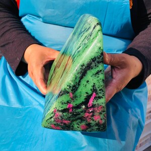 May include: A polished, triangular-shaped zoisite mineral specimen featuring a vibrant green base color with prominent black mineral inclusions and scattered, bright pink ruby crystals embedded throughout the smooth, reflective stone surface.