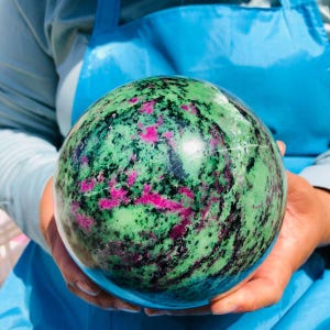 May include: A polished spherical stone featuring a vibrant mix of light green, deep magenta, and black mineral inclusions. The surface is smooth and reflective, showcasing a natural marbled pattern throughout the entire round mineral specimen.