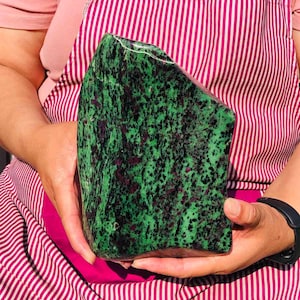 May include: A large, irregularly shaped slab of zoisite mineral featuring a vibrant green base color with prominent, scattered dark black hornblende inclusions and deep magenta-colored ruby crystal patches throughout the polished stone surface.