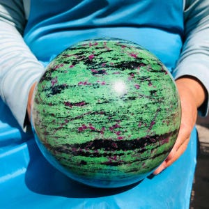 May include: A polished spherical stone featuring a vibrant green base color with prominent black streaks and scattered magenta-colored inclusions throughout the surface. The sphere displays a smooth, reflective texture with natural mineral patterns.